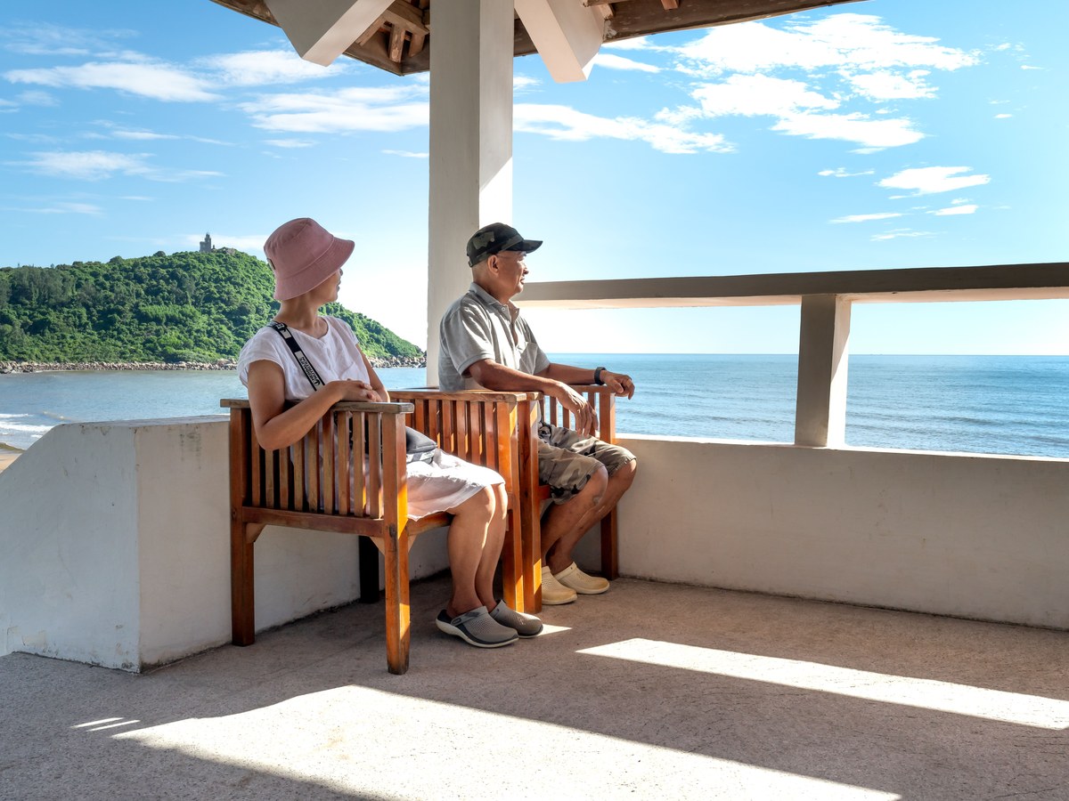 An older couple sitting on a shaded terrace bench, looking out over a calm bay
