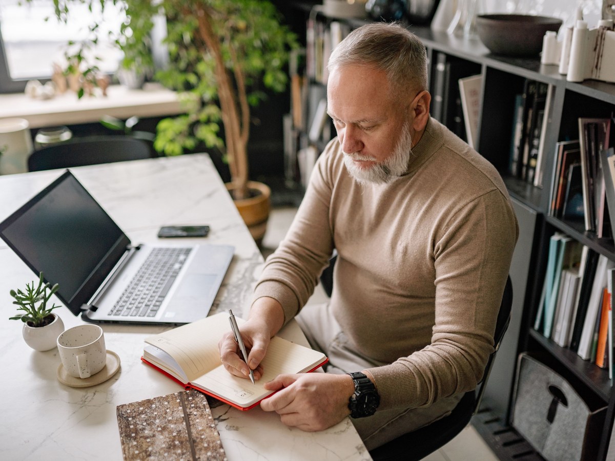 A man writing in a notebook at a desk beside an open laptop, working through the details of a trip